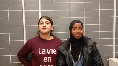 Två tjejer står uppställda mot en vägg och ser självsäkra ut.
